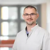 Portret mężczyzny w okularach i białym fartuchu lekarskim, uśmiechniętego.