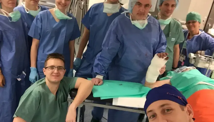Zespół chirurgów w strojach operacyjnych pozuje do zdjęcia w sali operacyjnej, jeden z nich trzyma nogę w gipsie.